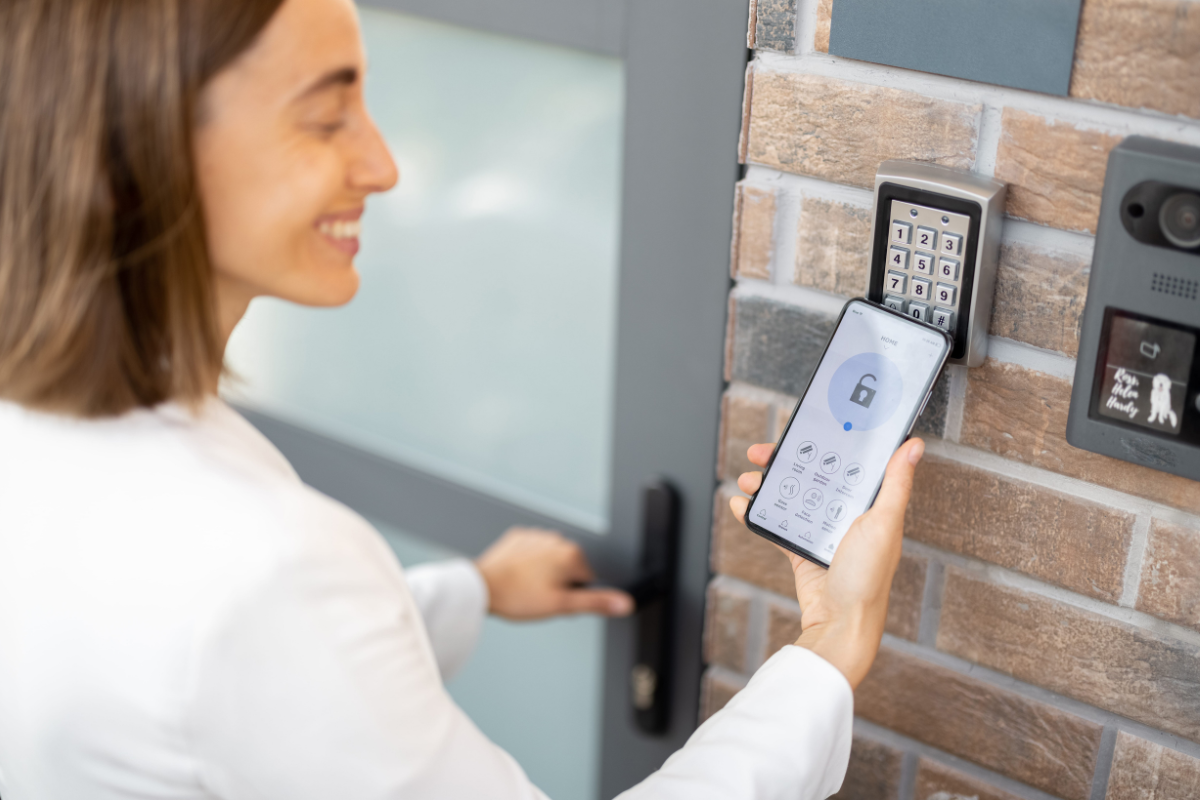 Person using a smartphone near a keypad smart lock mounted on a brick wall at an entry door
