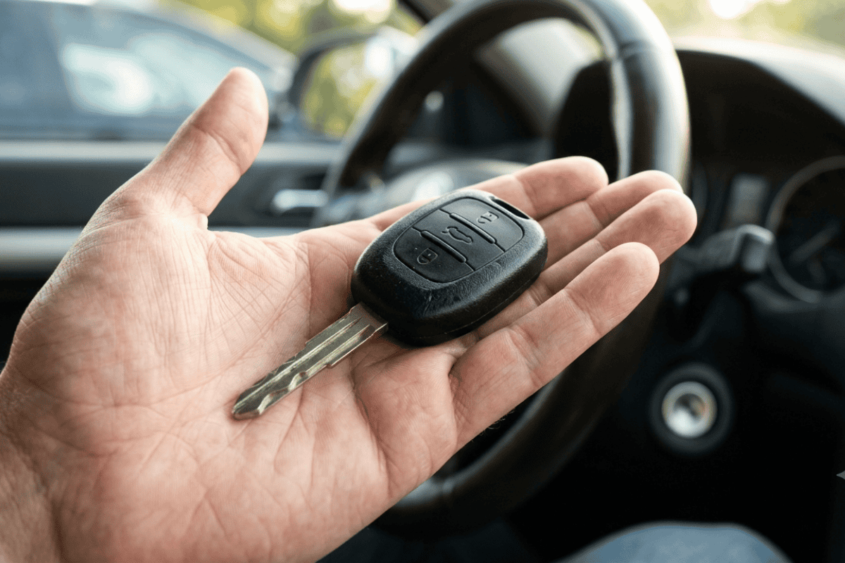 Hand holding a car key fob with three buttons, resting over a car steering wheel interior visible in the background.